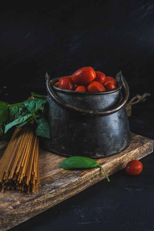 Free A rustic food arrangement featuring cherry tomatoes in a pot with spaghetti and basil leaves on a wooden board. Stock Photo
