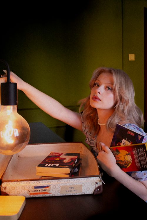 Free A young woman holding books near a warm light in a cozy room, creating a soft and artistic feel. Stock Photo