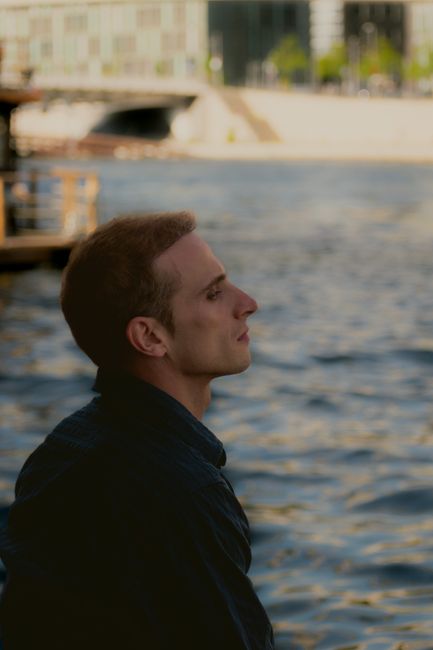 A man with short hair stands by an urban river, deep in thought.