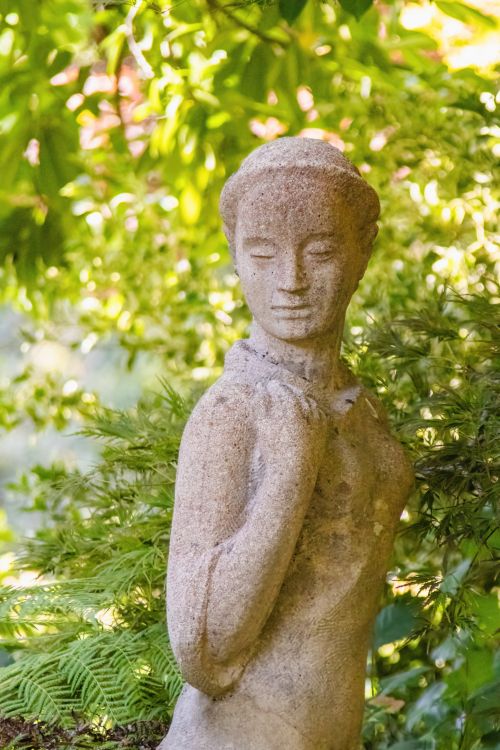 Free Elegant stone statue surrounded by lush greenery in a sunlit garden. Stock Photo