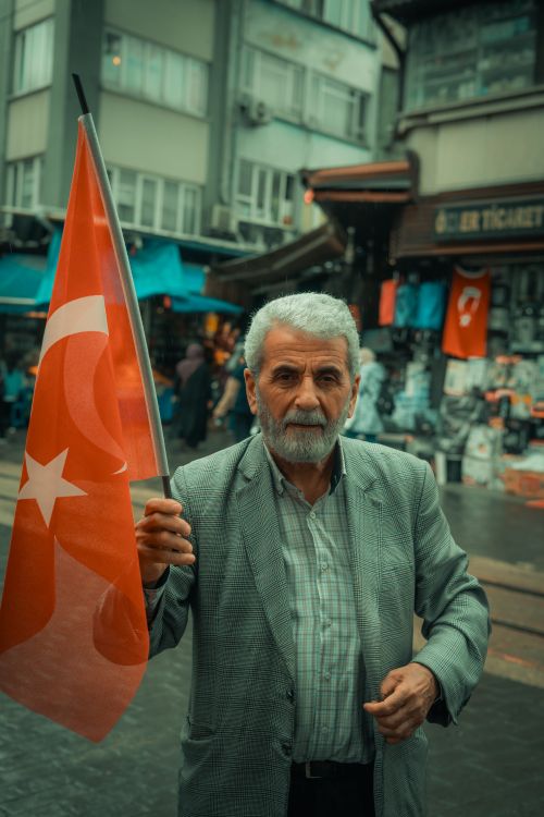 Free Elderly man proudly holding a Turkish flag in a vibrant Bursa street market. Stock Photo