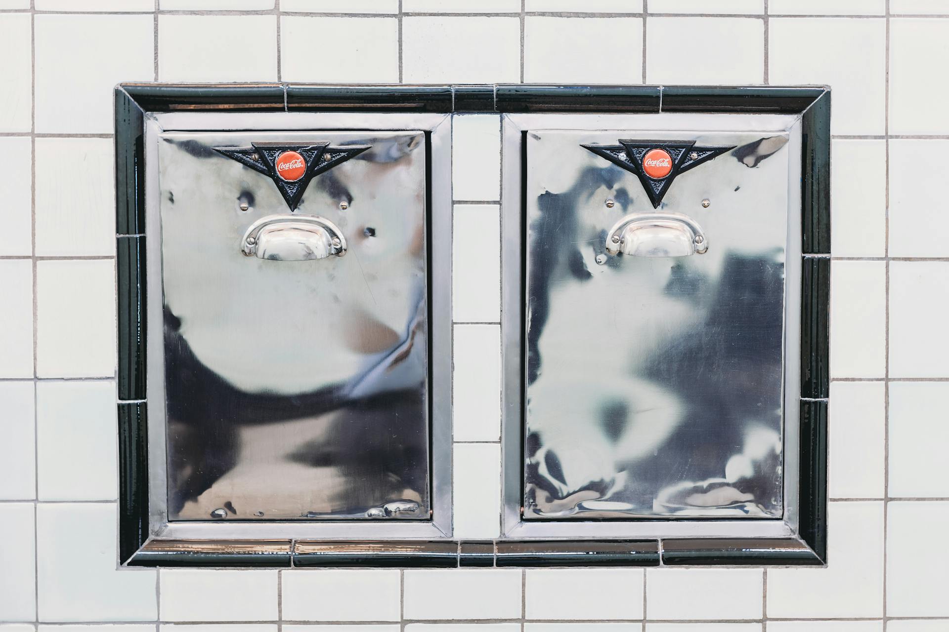 Close-up of vintage Coca-Cola cooler doors with handles on a tiled wall.
