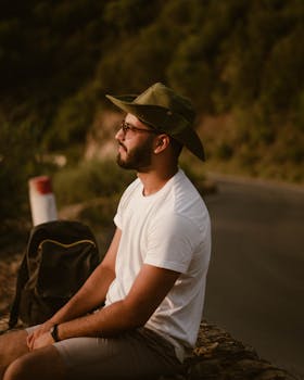 Man in a hat sitting by the road with a backpack during sunset, captured in a serene outdoor setting.