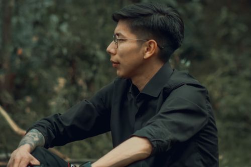 Free Thoughtful man with glasses and tattoos sitting in a park, wearing a black shirt. Stock Photo