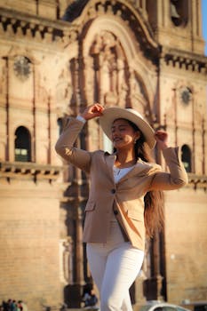 Woman in tan jacket and white pants smiling joyfully near historic architecture under a sunny sky.