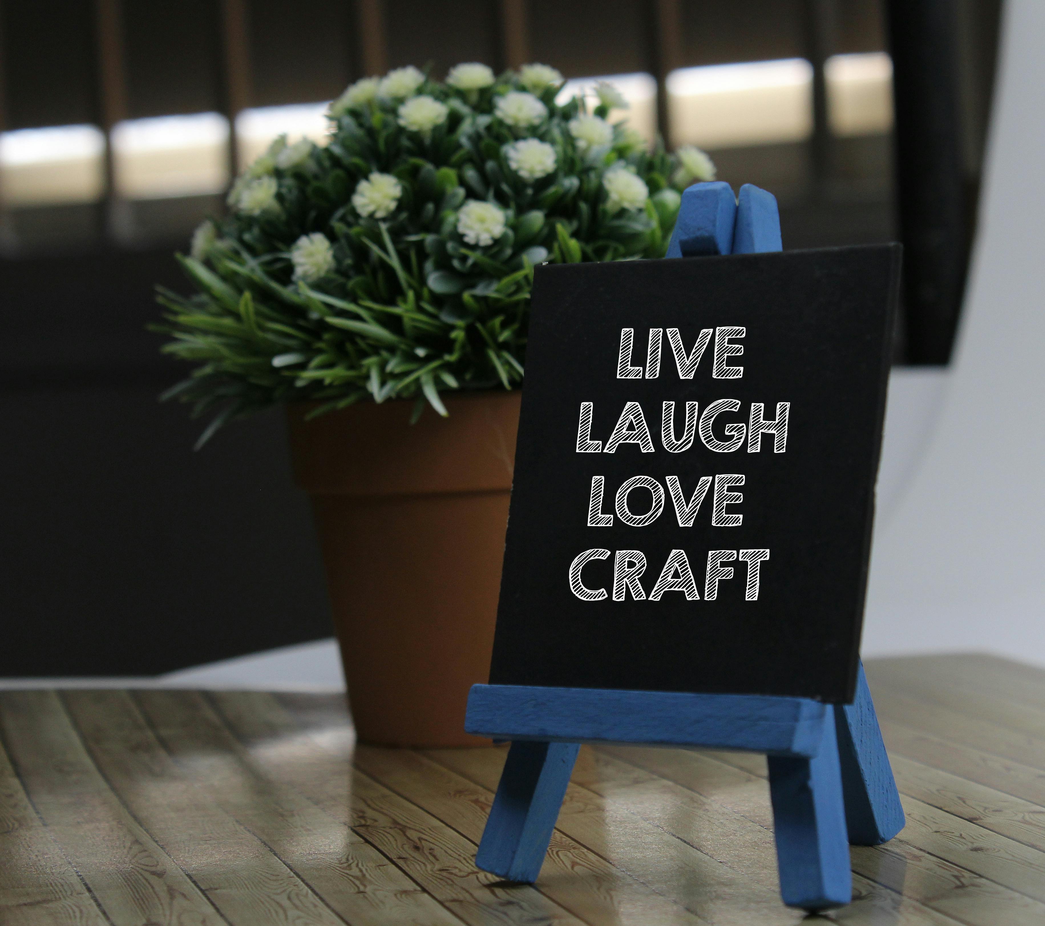 Chalkboard on an easel with 'Live Laugh Love Craft' text beside a plant in a pot.