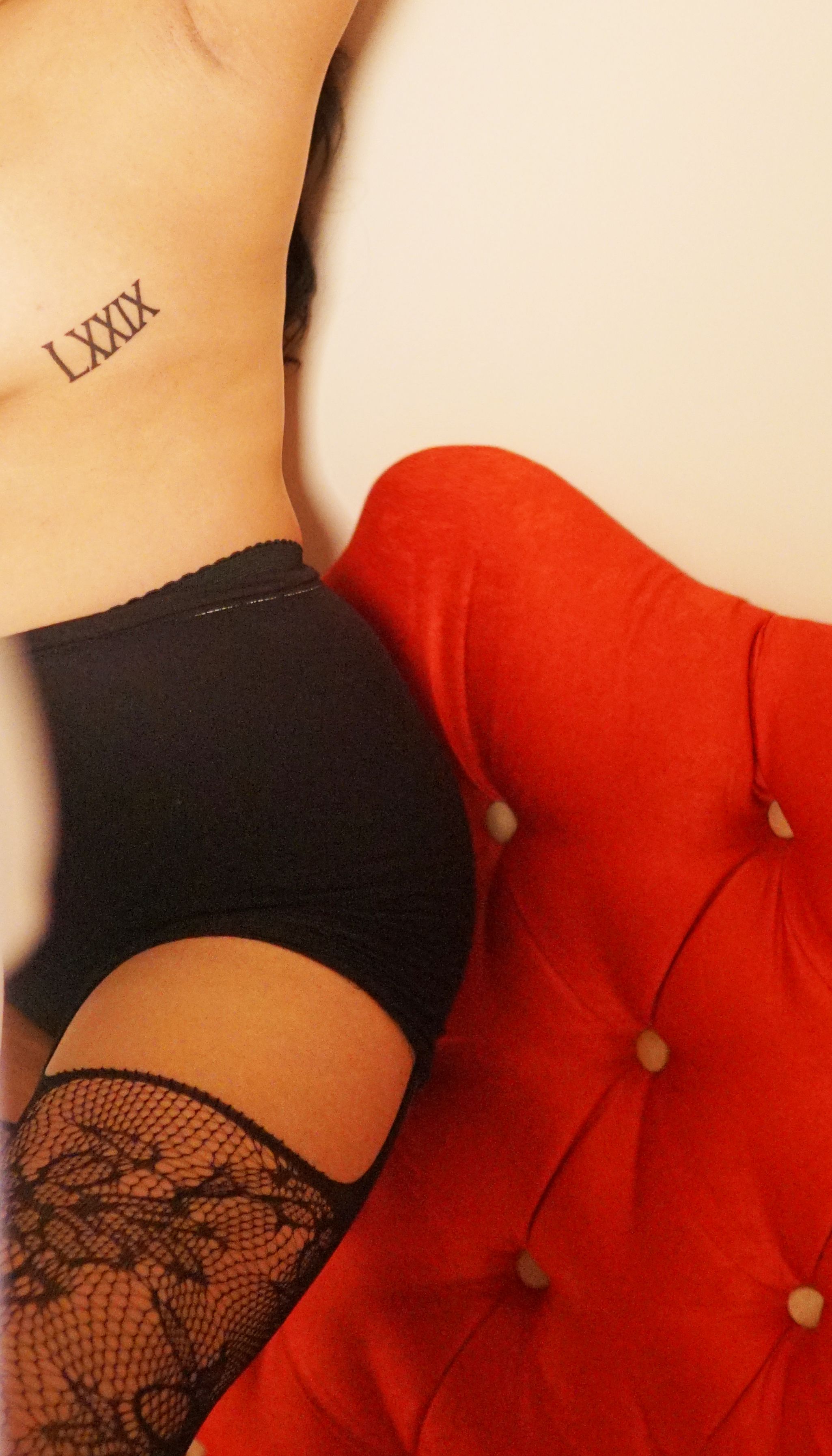 Artistic nude captures seduction with tattooed figure and lace stockings on a red armchair.