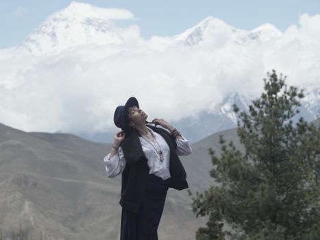 A woman in a hat poses beautifully in a picturesque mountain setting with trees and clouds.