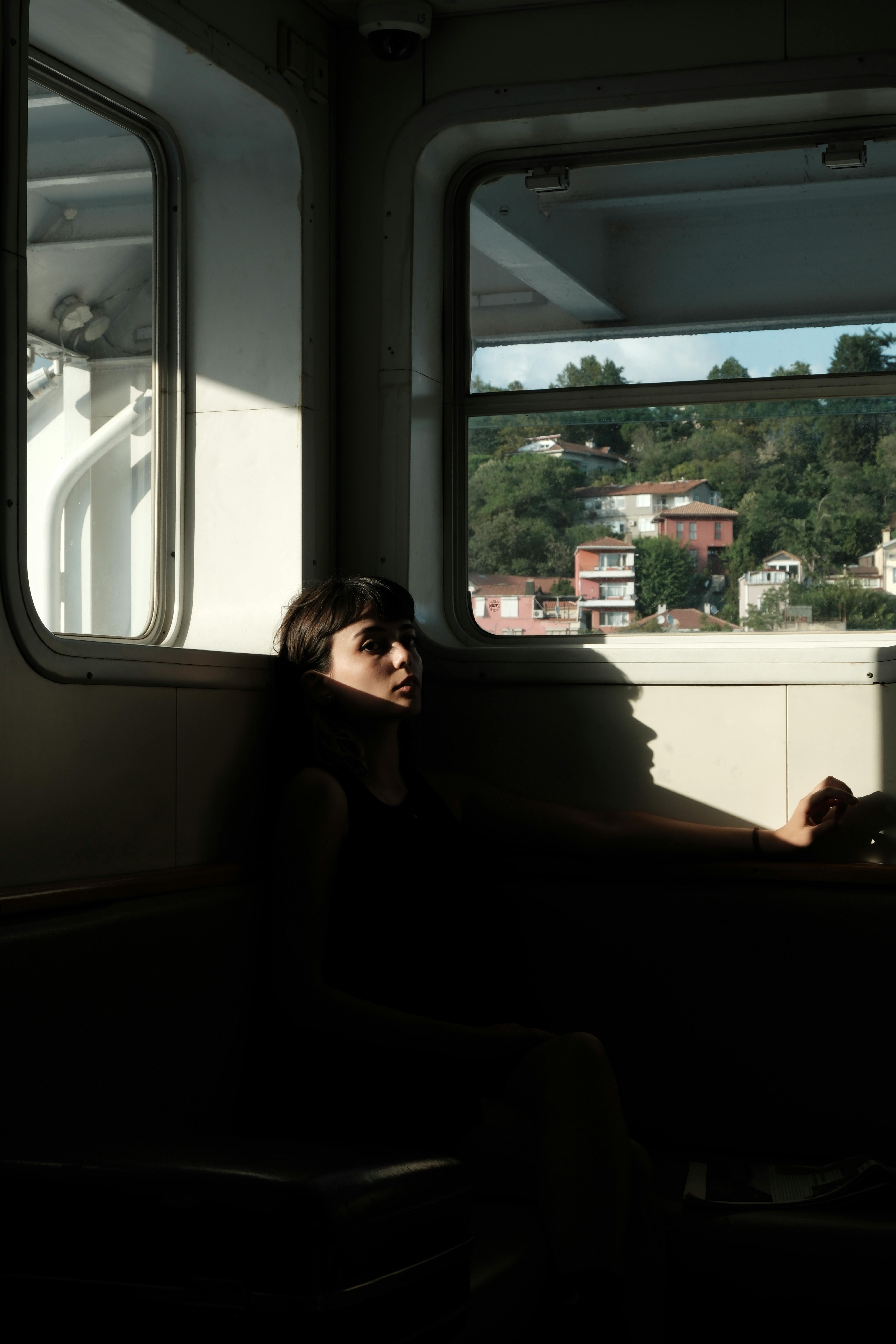 A woman sits quietly inside a ferry, with scenic views through large windows.