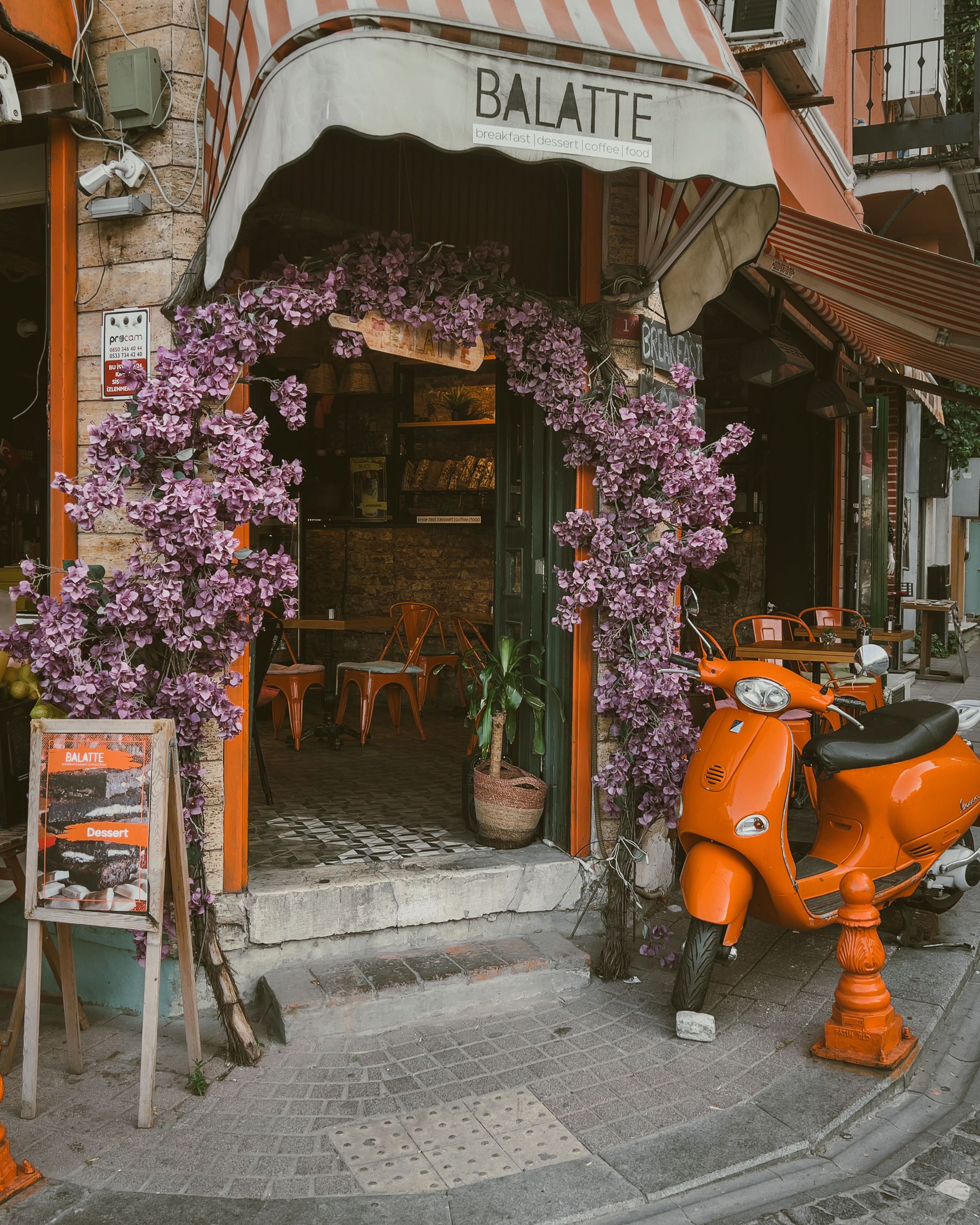 A quaint café adorned with flowers and an orange scooter parked outside, creating a vintage urban vibe.