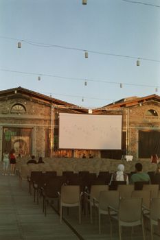 An outdoor cinema setup with empty seats facing a large screen under a clear sky at sunset.