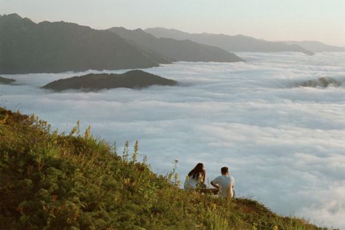 Free A couple enjoys a serene view of mountains and clouds from a grassy hilltop. Stock Photo