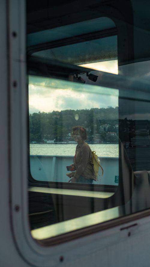 Free A woman stands on a ferry deck, visible through a ship window with water and trees in the background. Stock Photo