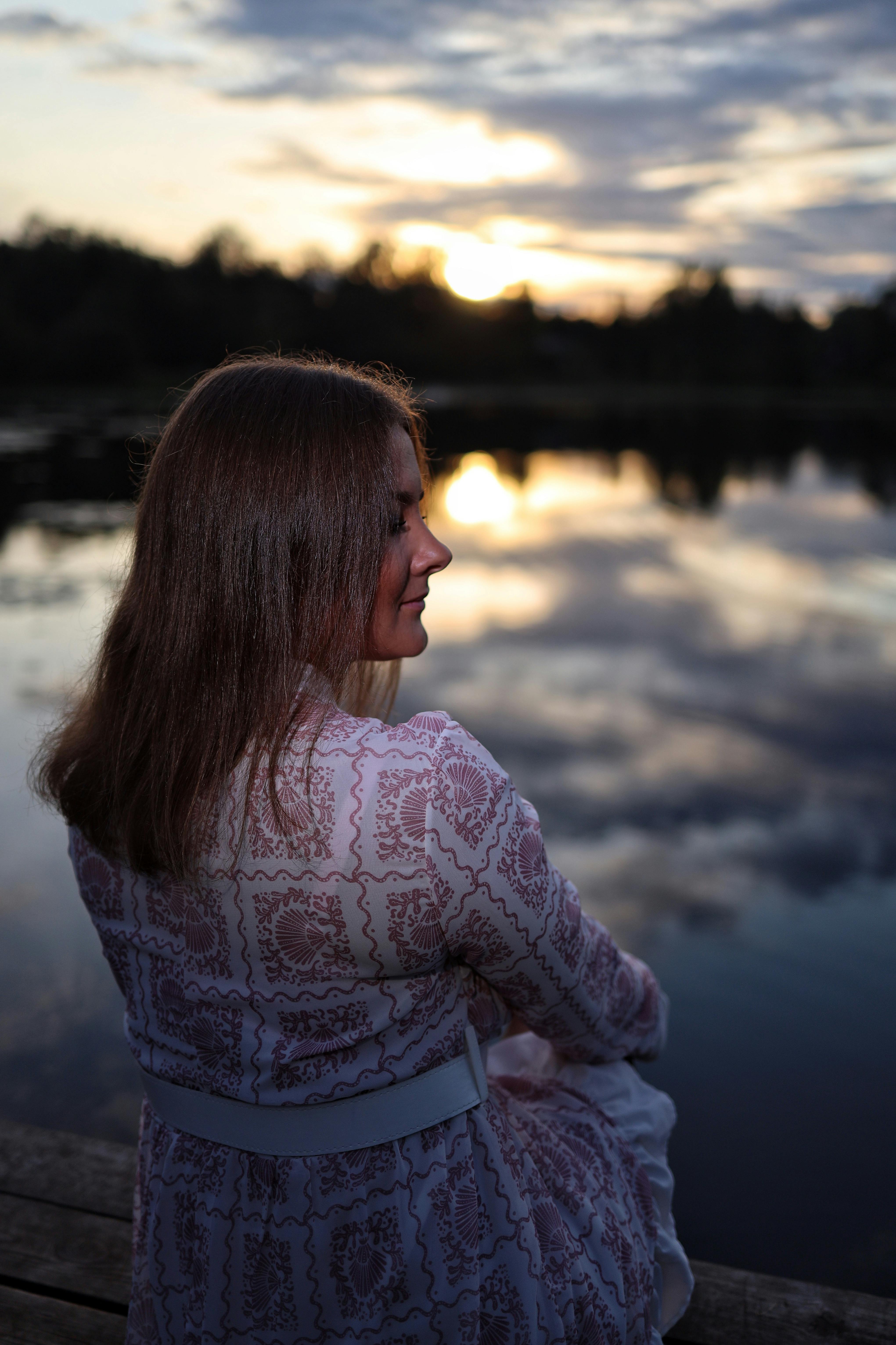 A woman in a patterned dress sits by the serene lake at sunset, reflecting tranquility.