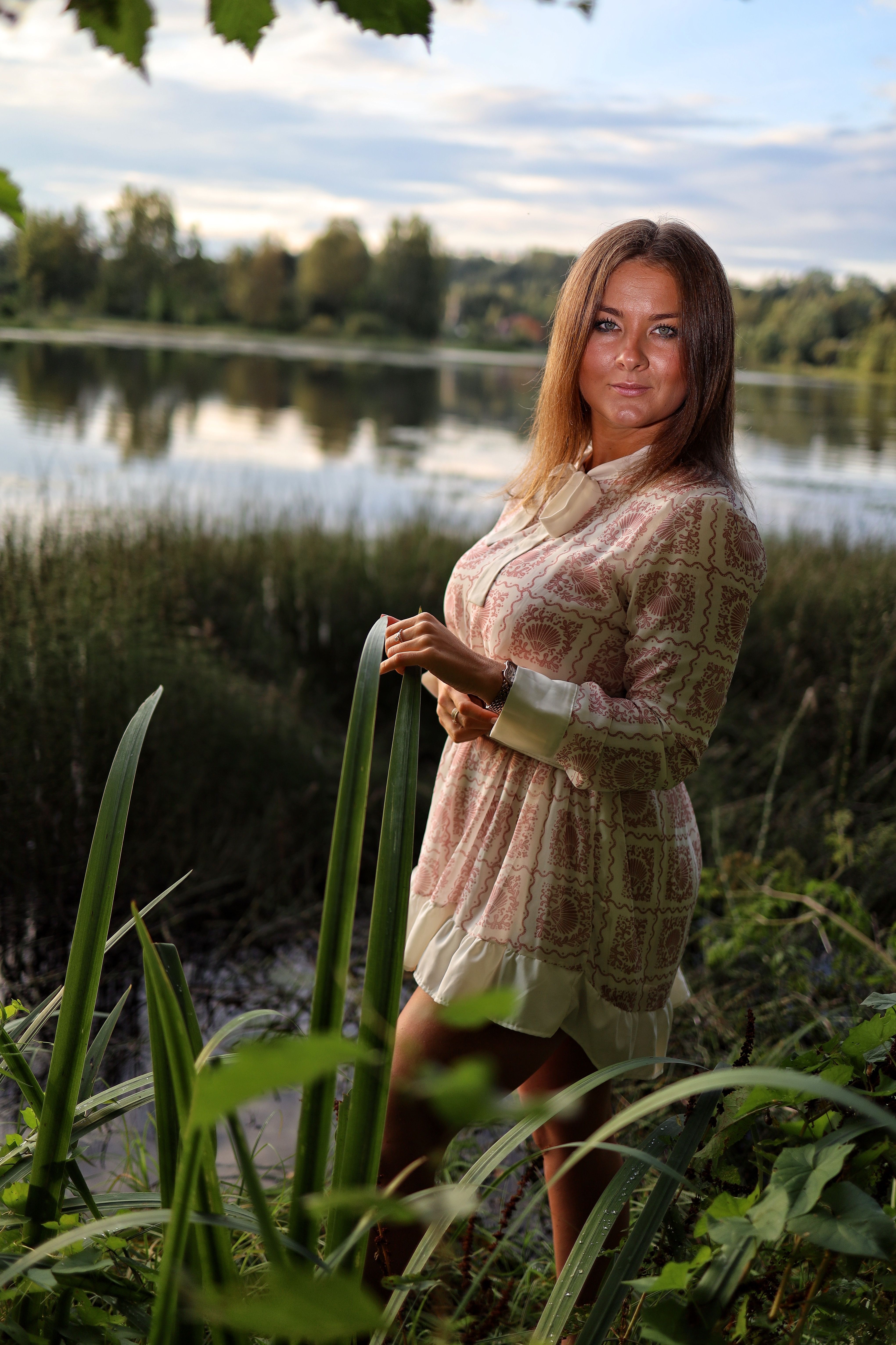 A woman in a summer dress stands by a serene lake surrounded by lush greenery.