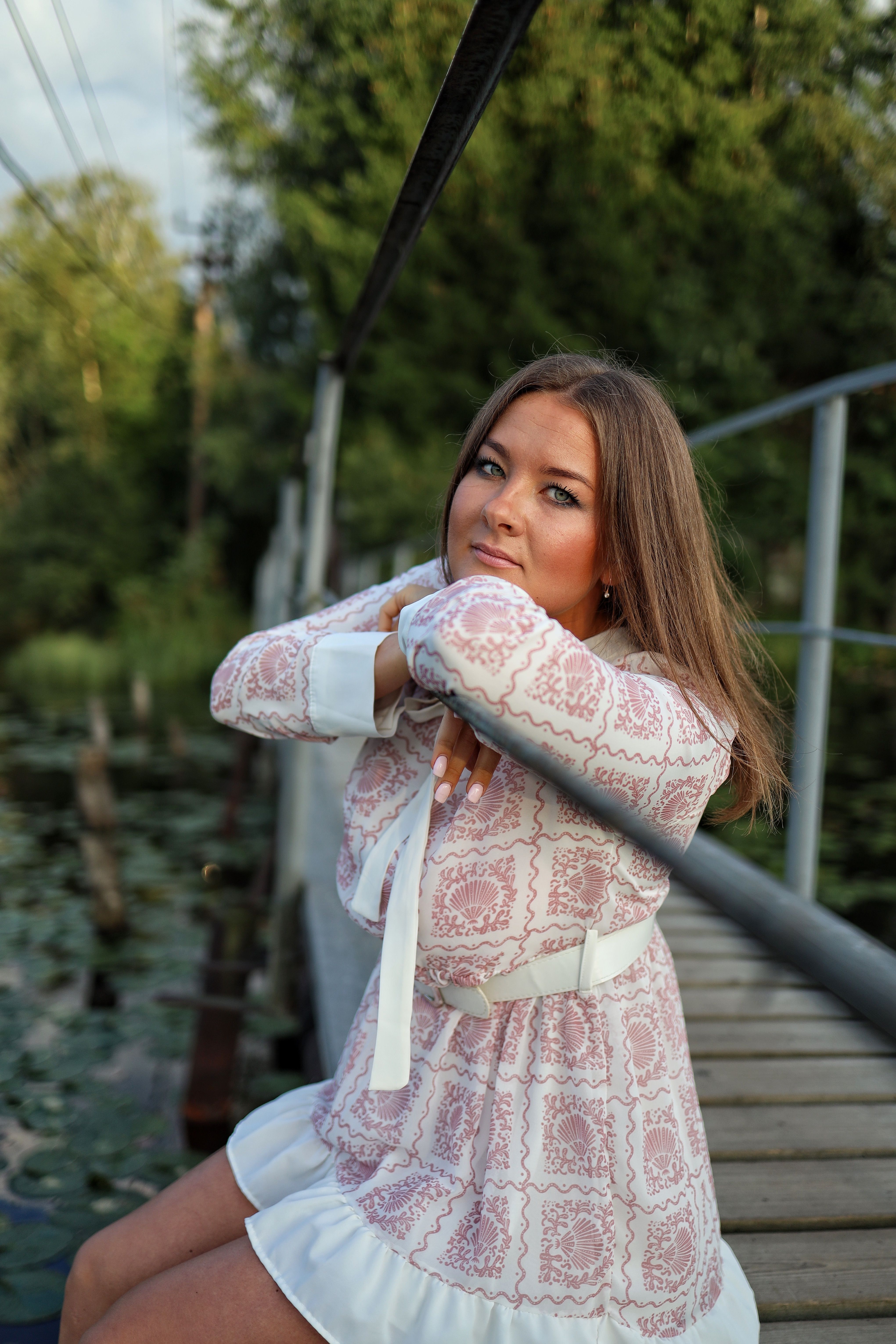 Young woman in a patterned dress poses on a serene wooden footbridge in a summer park.