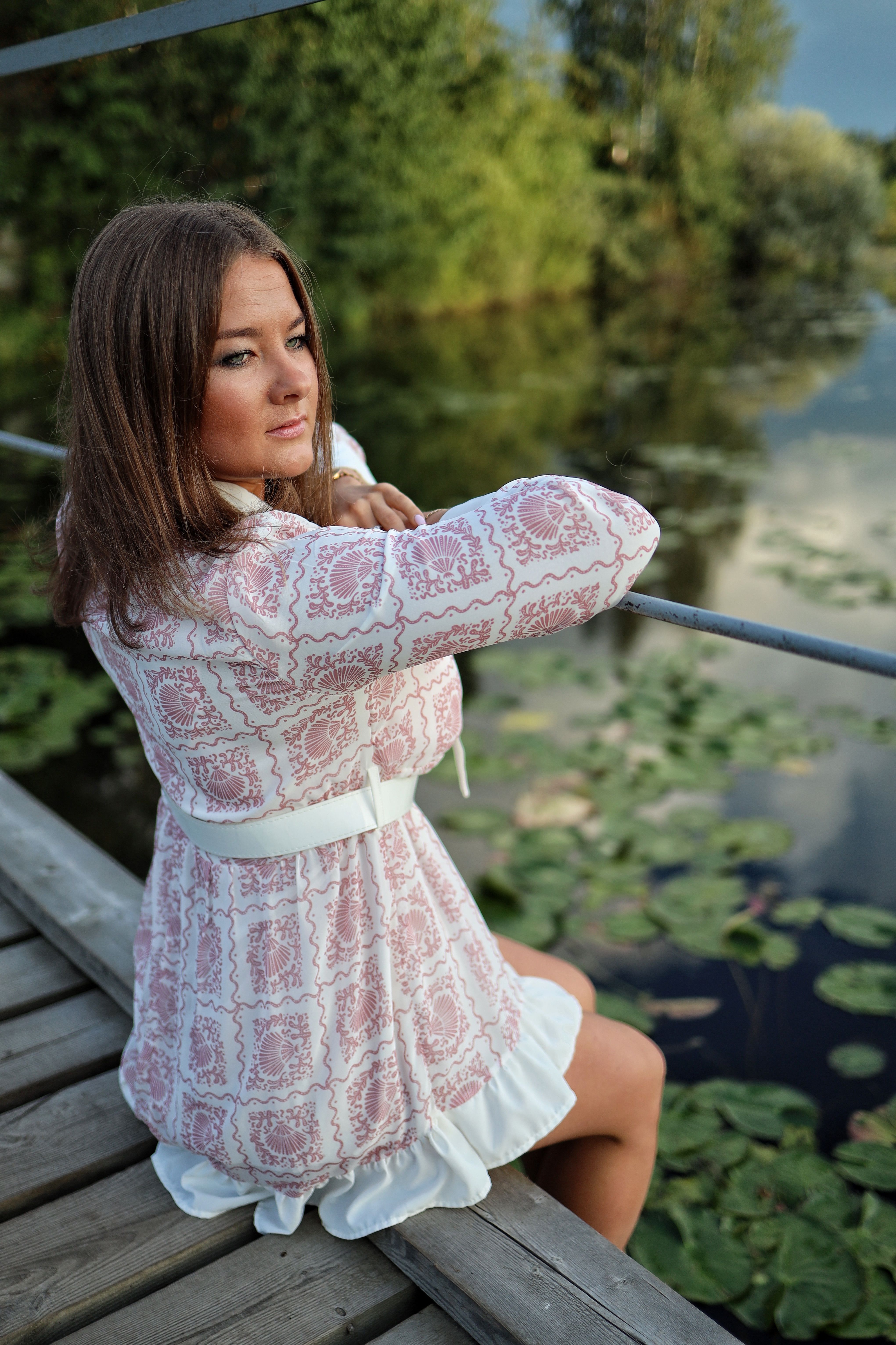 A serene scene of a woman in a patterned dress sitting on a wooden footbridge, overlooking a tranquil pond with lily pads.
