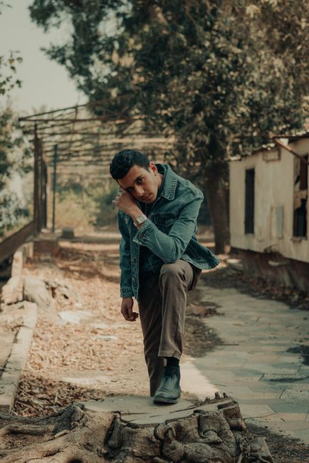 Man in a denim jacket poses confidently outdoors amid trees and rustic structures.