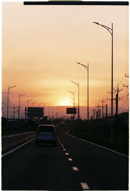 A serene shot of a car driving on an empty road during sunset, highlighted by streetlights and soft hues.
