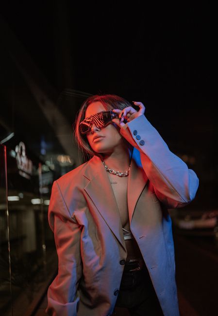 Young woman in a trendy blazer posing with sunglasses under neon lights in an urban setting.