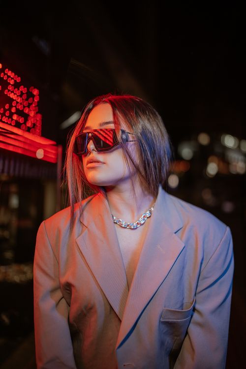 Free Young woman in fashionable blazer and sunglasses posing under urban neon lights at night. Stock Photo