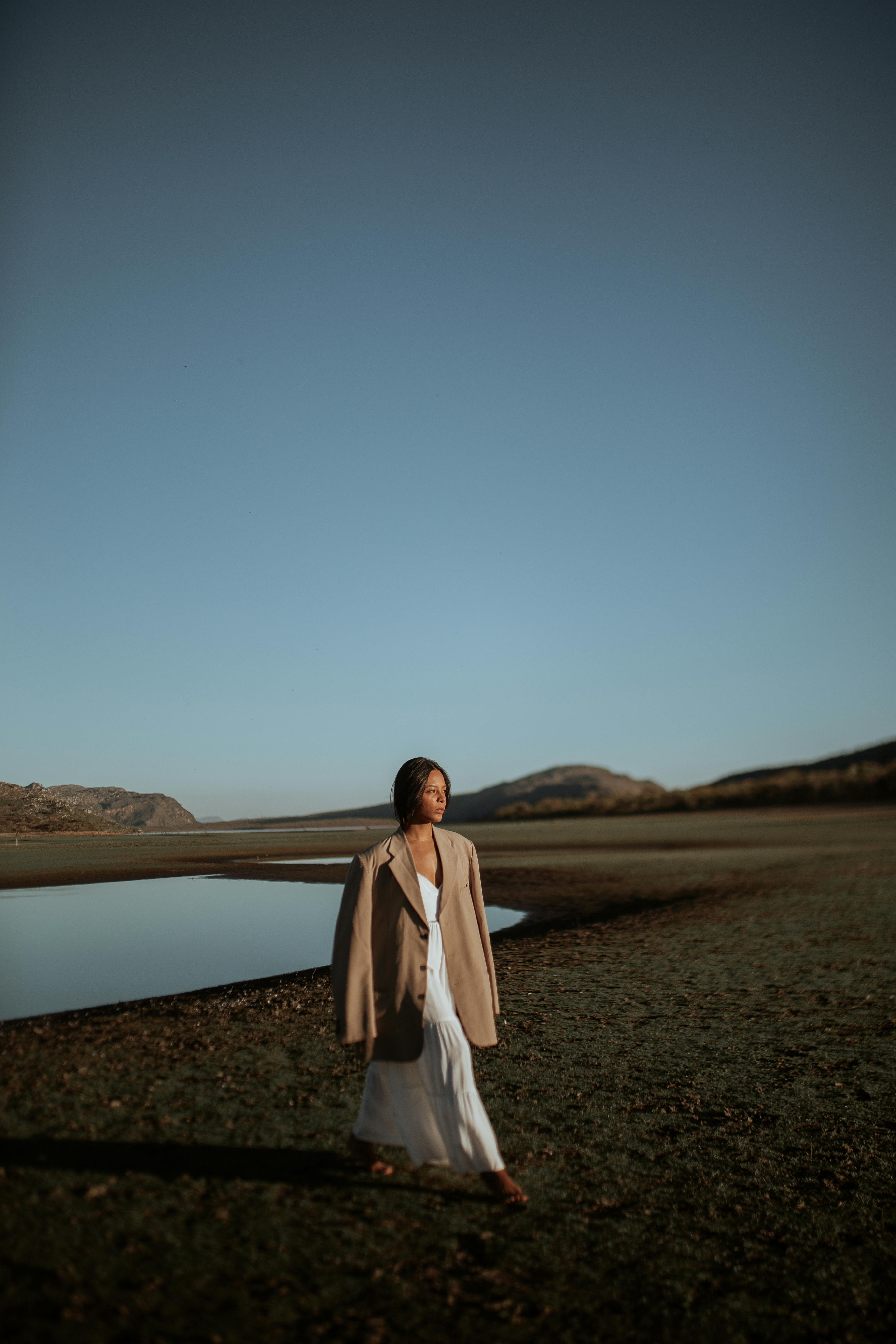 Fashionable woman in a blazer walks by a tranquil lakeside with mountains in the background.