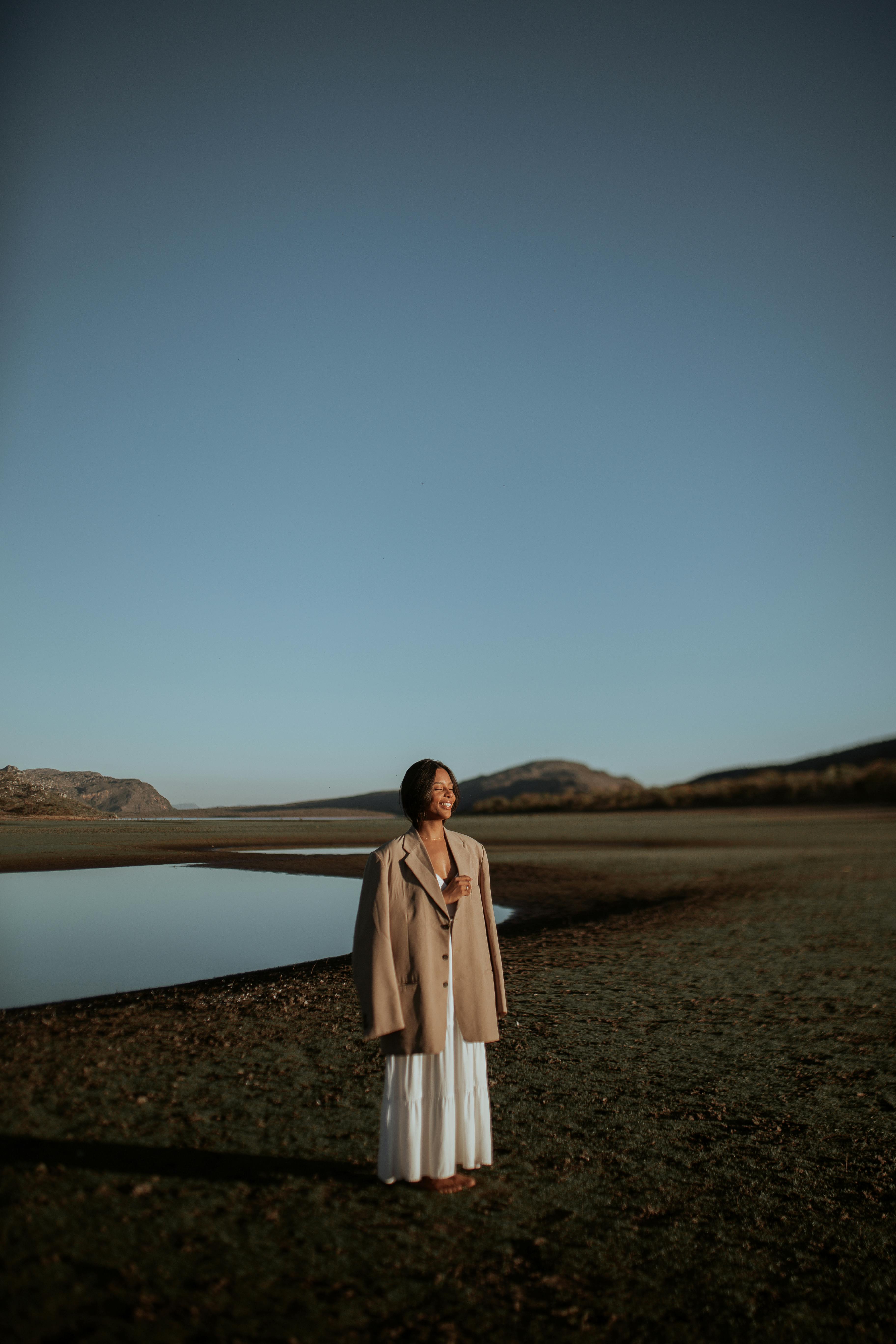 A woman in a beige coat stands peacefully in a vast, rural landscape under a clear blue sky.