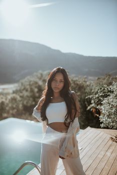 Fashionable young woman in white crop top and beige pants posing by sunlit poolside outdoors.
