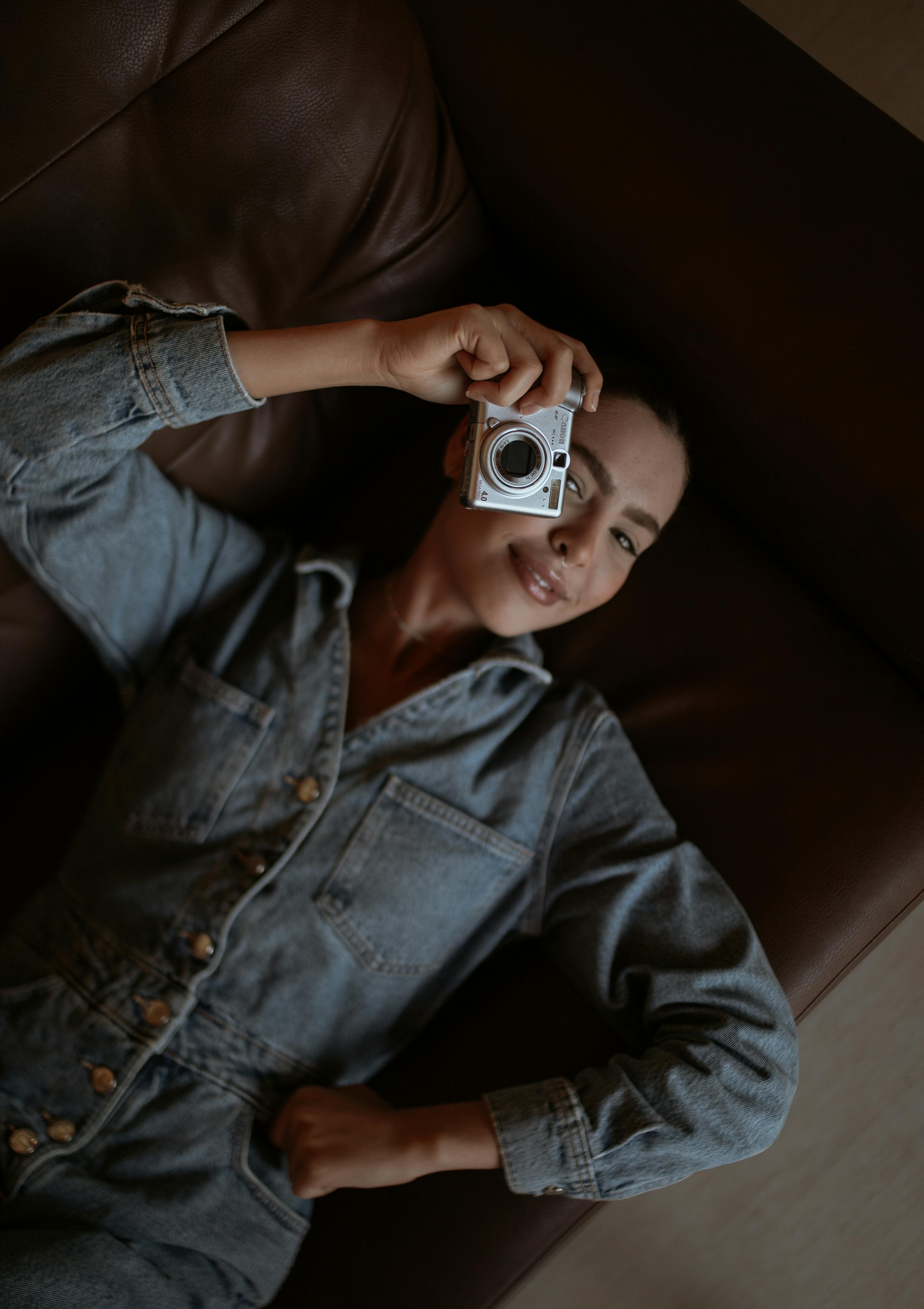 Young woman in denim overalls holding a camera while lying on a leather couch.