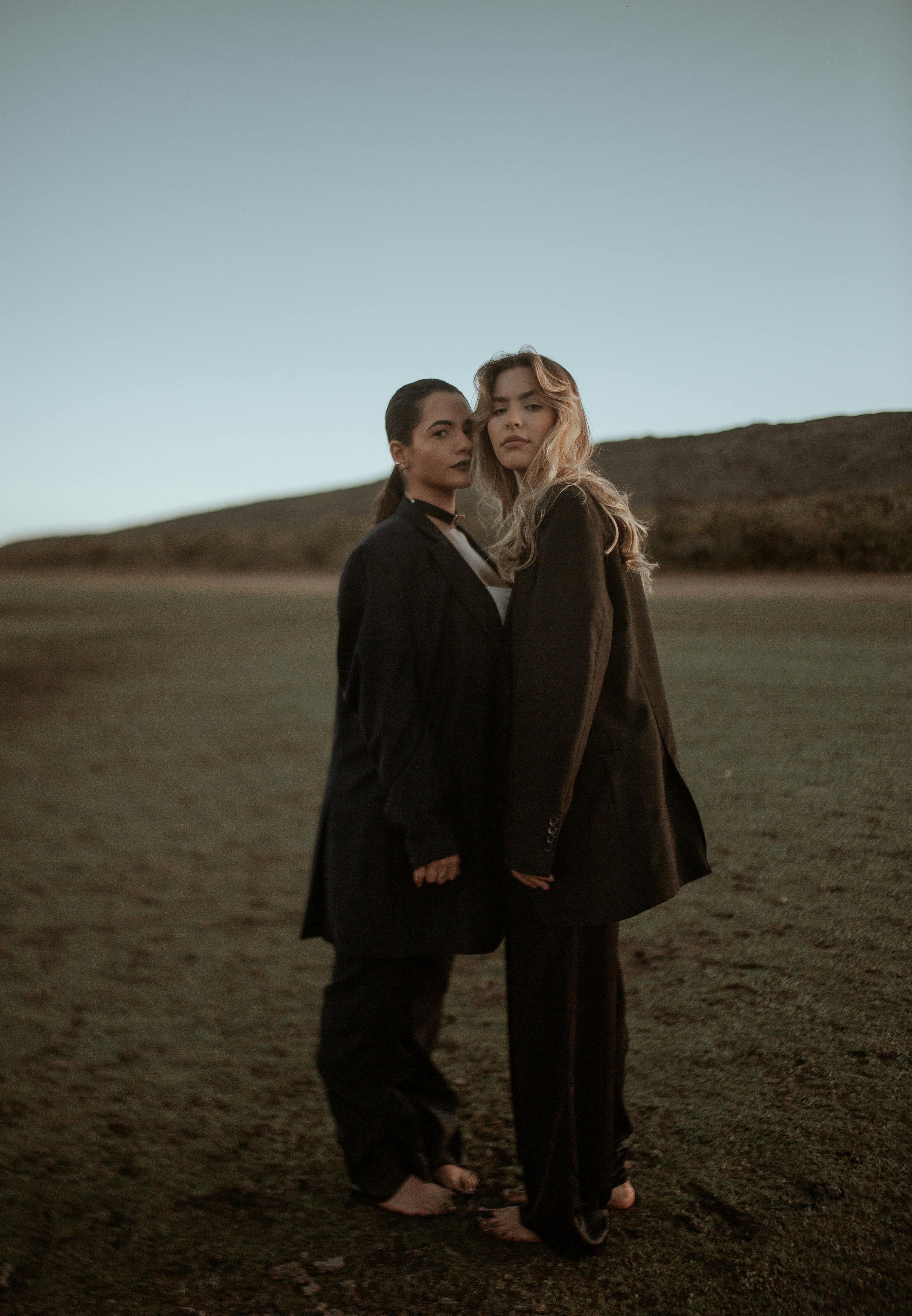 Two women in elegant suits pose barefoot on a grassy field, showcasing contemporary fashion.
