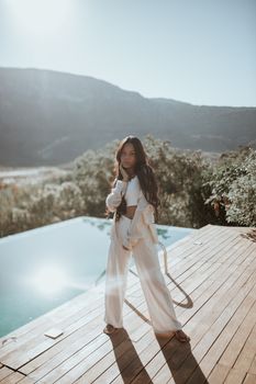 Fashionable woman in white attire poses by a pool with scenic mountain backdrop.