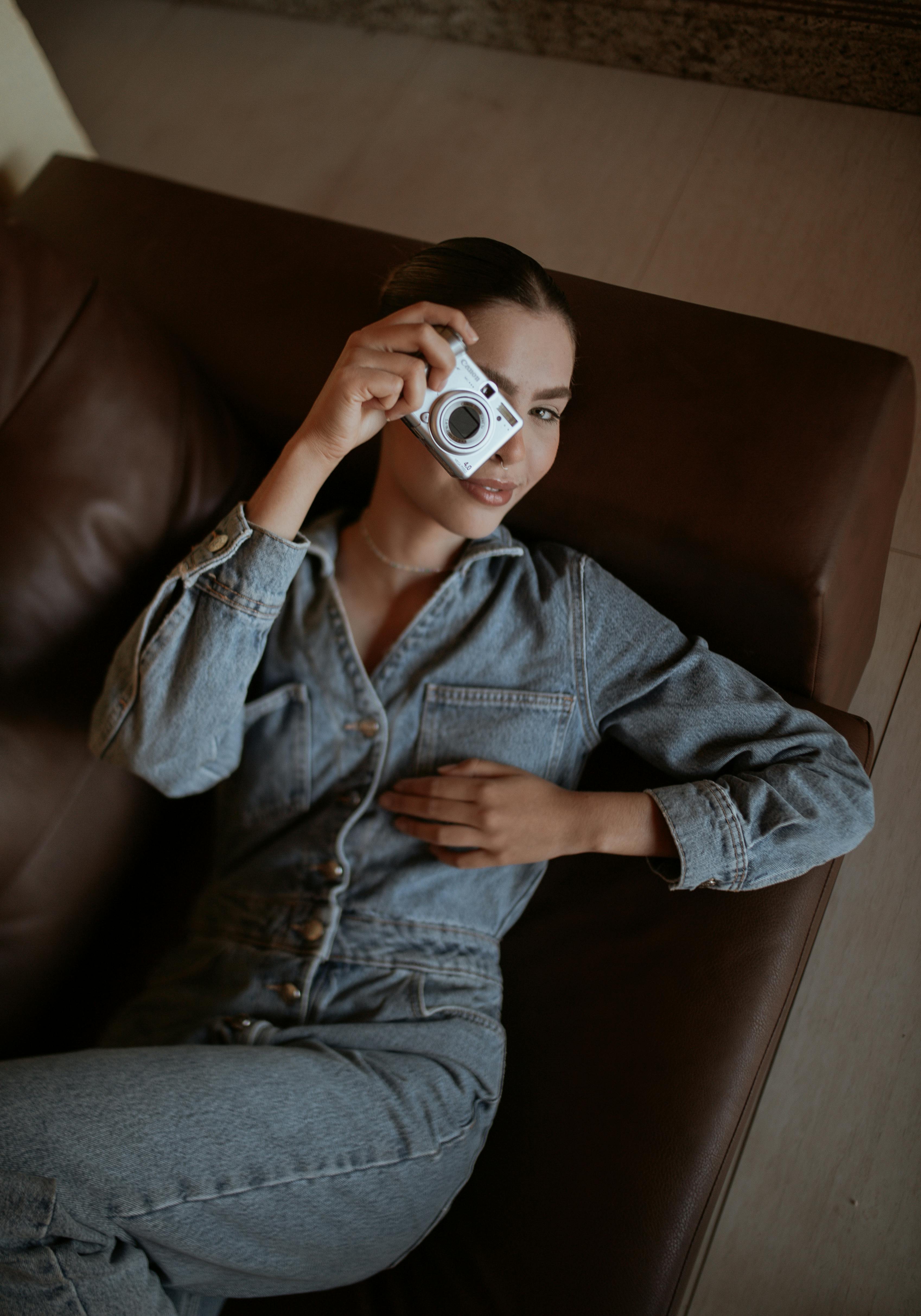 A stylish woman in denim holds a camera while lounging on a sofa indoors.