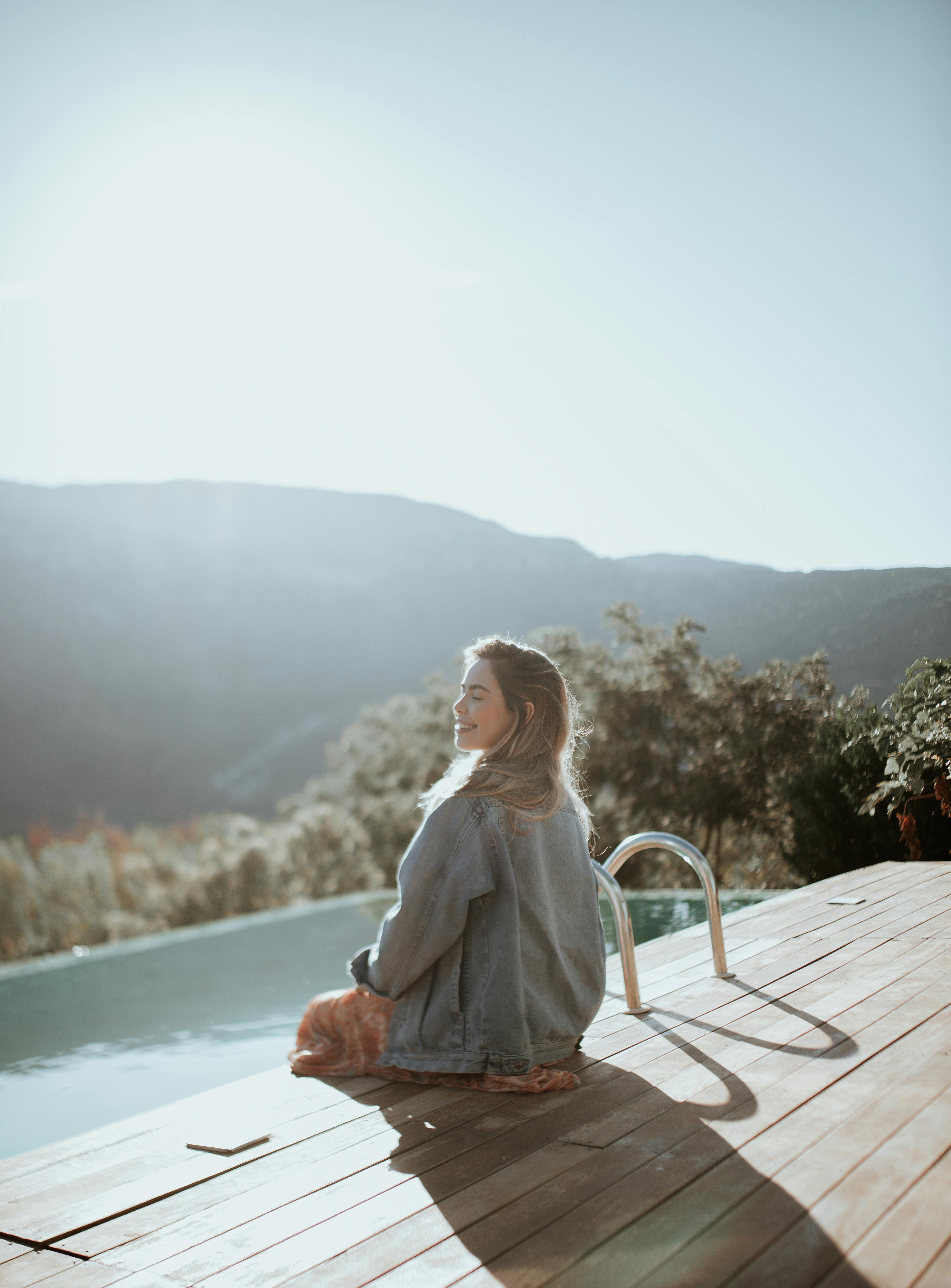 Woman enjoying a peaceful moment by an infinity pool with mountain views under a bright summer sky.