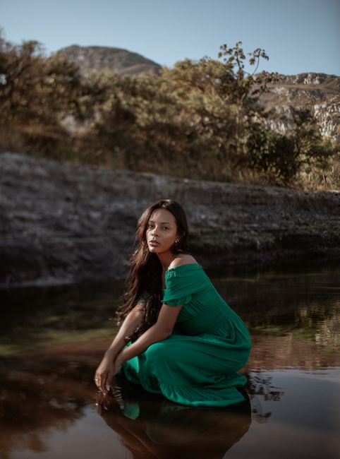 Young woman poses gracefully by a river, wearing a flowing green dress in a tranquil outdoor setting.