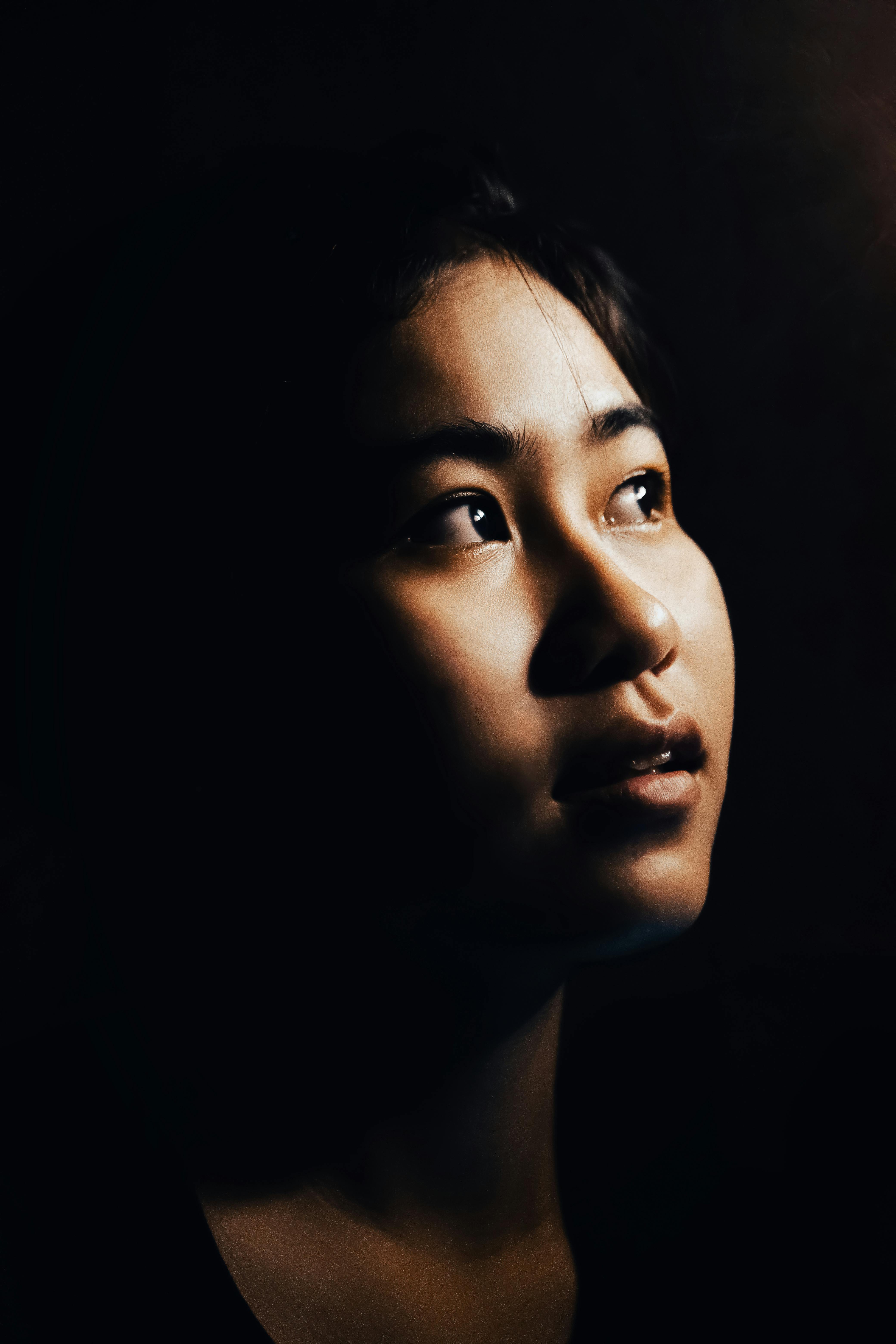 Portrait of an East Asian woman looking upwards with dramatic lighting on a black background.