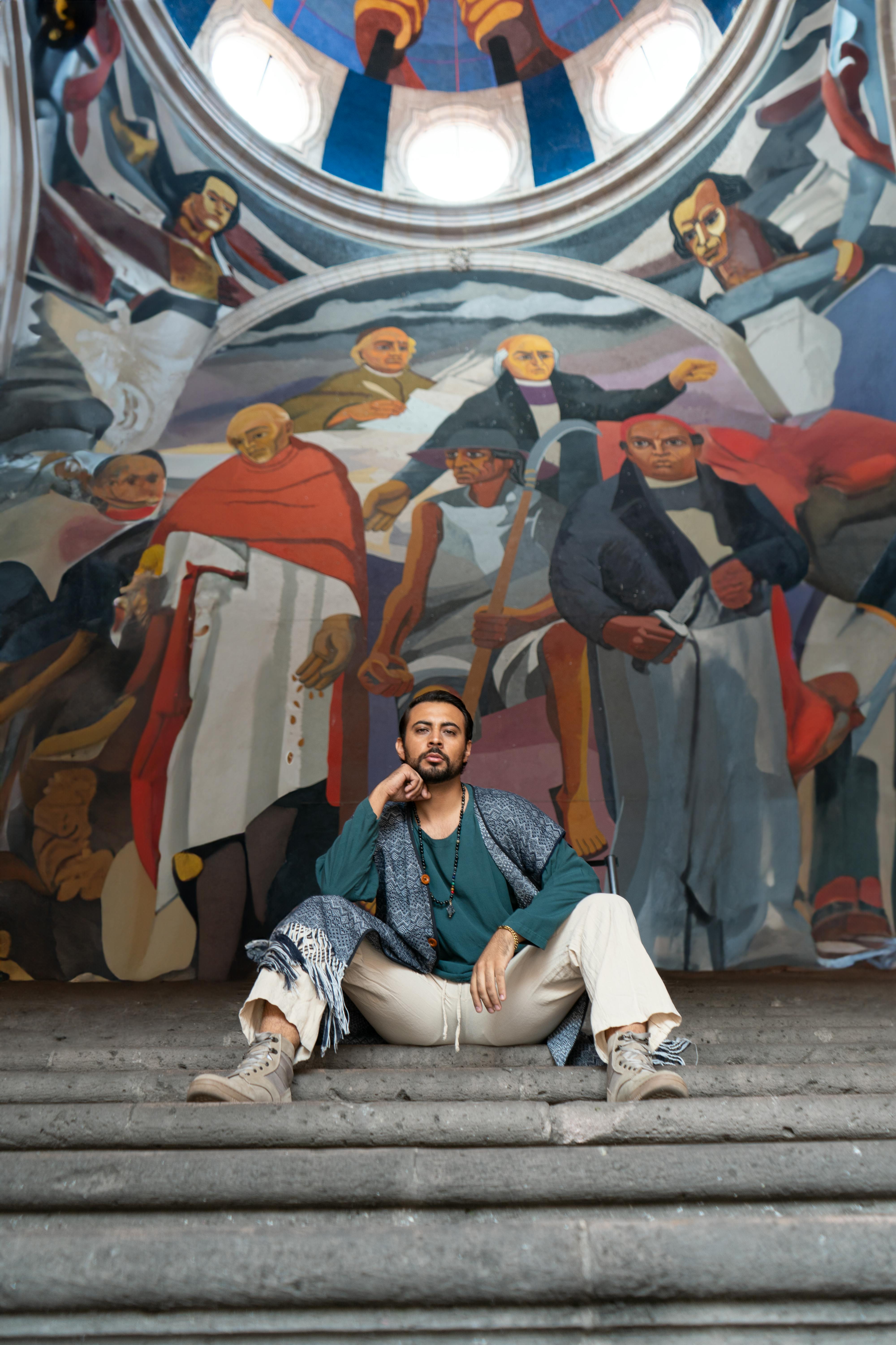 Stylish man sitting on stone steps with a colorful mural in Morelia, Mexico in the background.