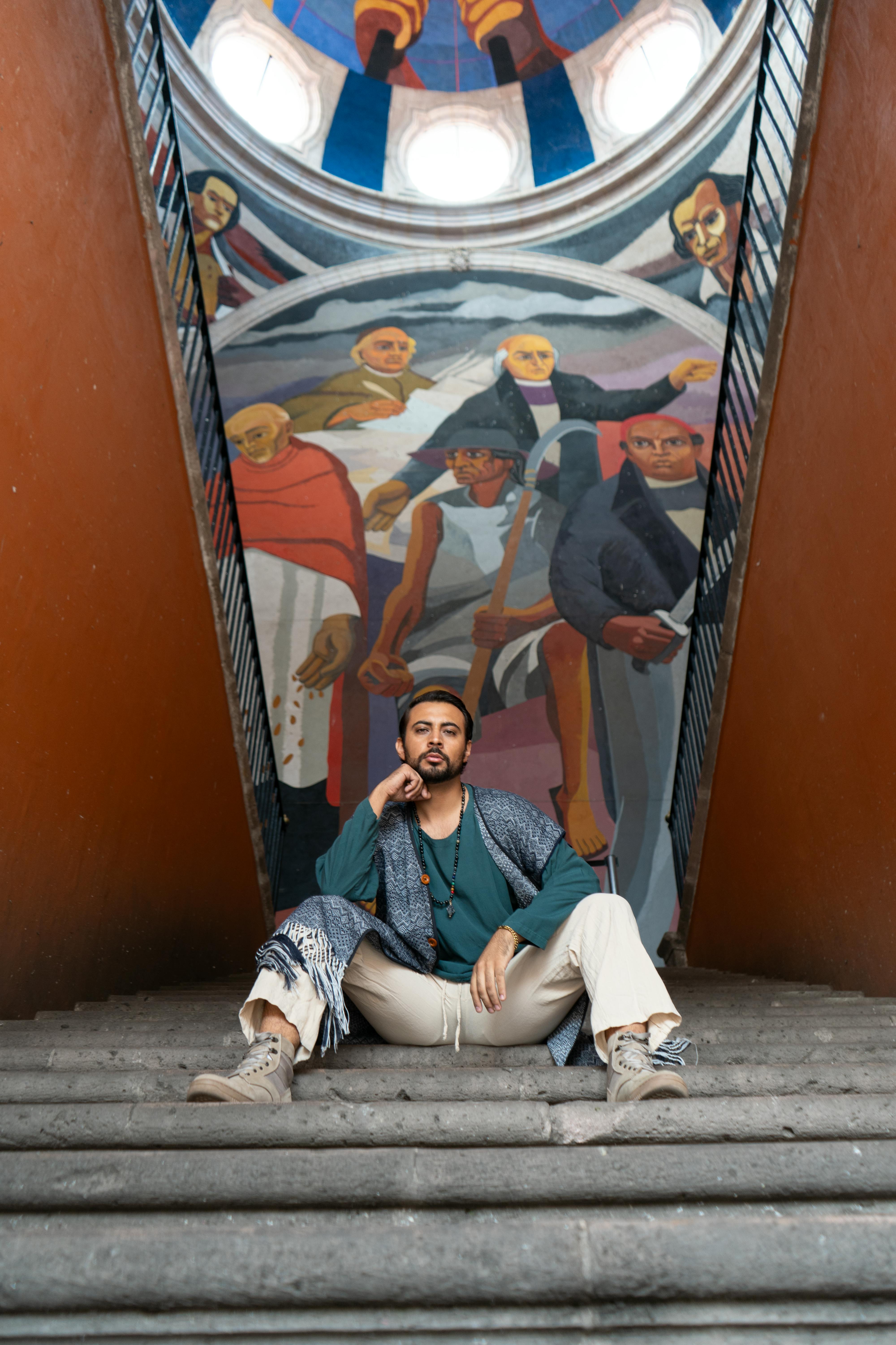 A man confidently sits on stairs in front of a colorful urban mural, indoors.