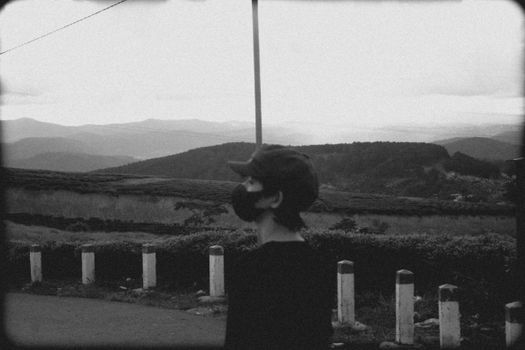 A solitary figure in a cap and mask gazes at the vast rural hills in black and white.
