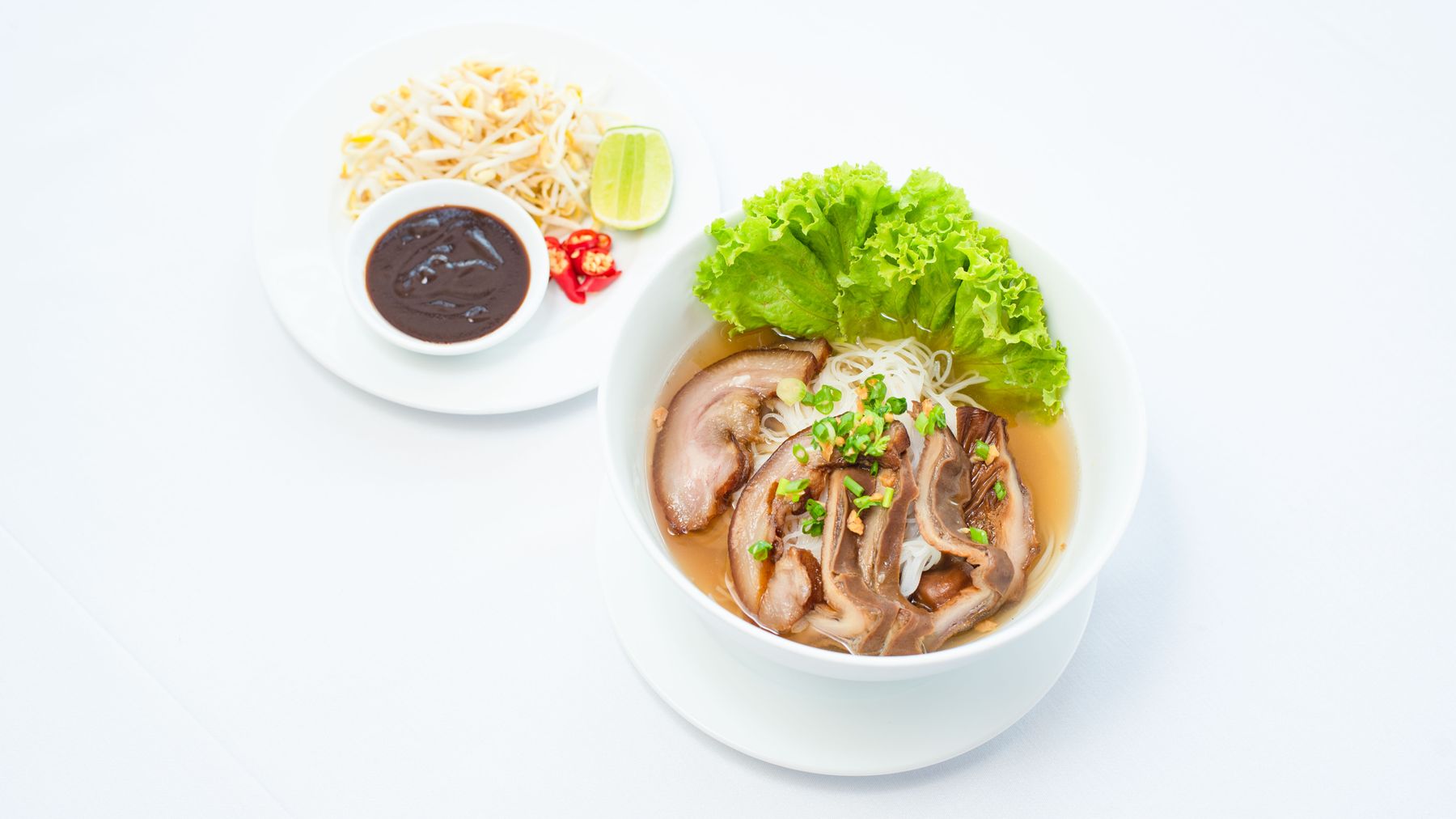A close-up of Asian pork noodle soup with lettuce, garnishes, and a side dish.