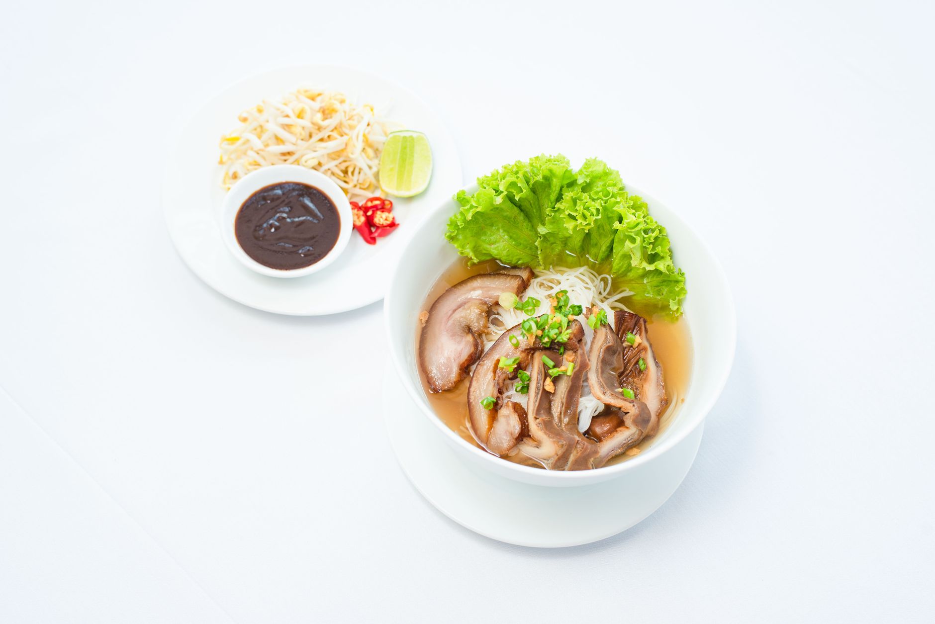 A close-up of Asian pork noodle soup with lettuce, garnishes, and a side dish.