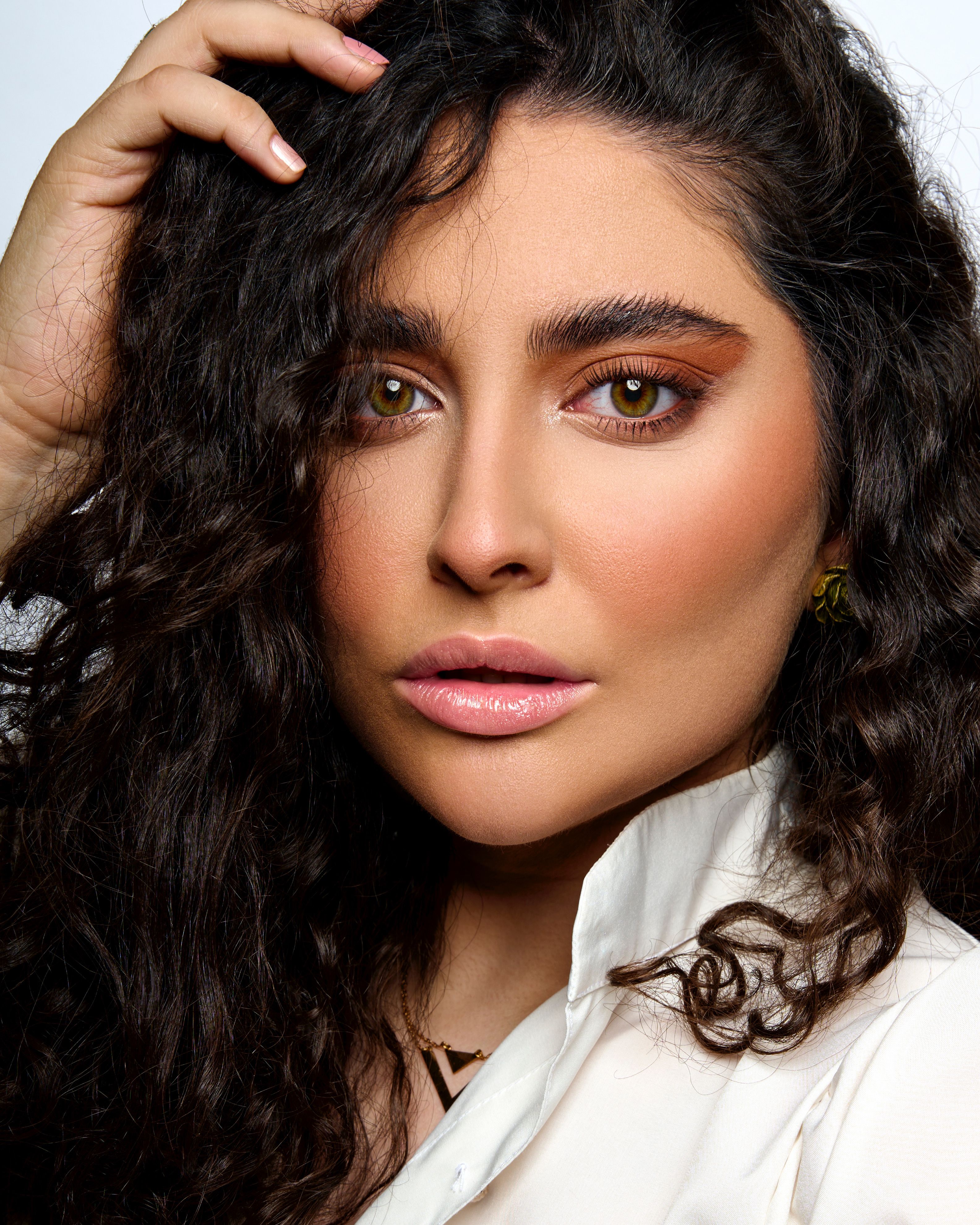 Close-up portrait of a woman touching her curly hair with dramatic makeup and confident expression.