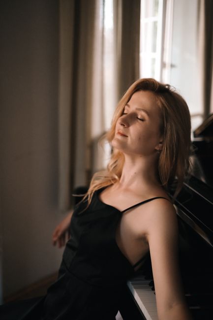 A blonde woman in a slip dress leans gracefully on a piano, exuding elegance and calm.
