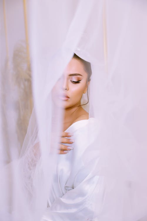 Young Woman in White Wedding Dress Posing Behind Tulle Curtains