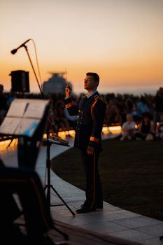 A conductor in military uniform leads an orchestra during a sunset performance outdoors.