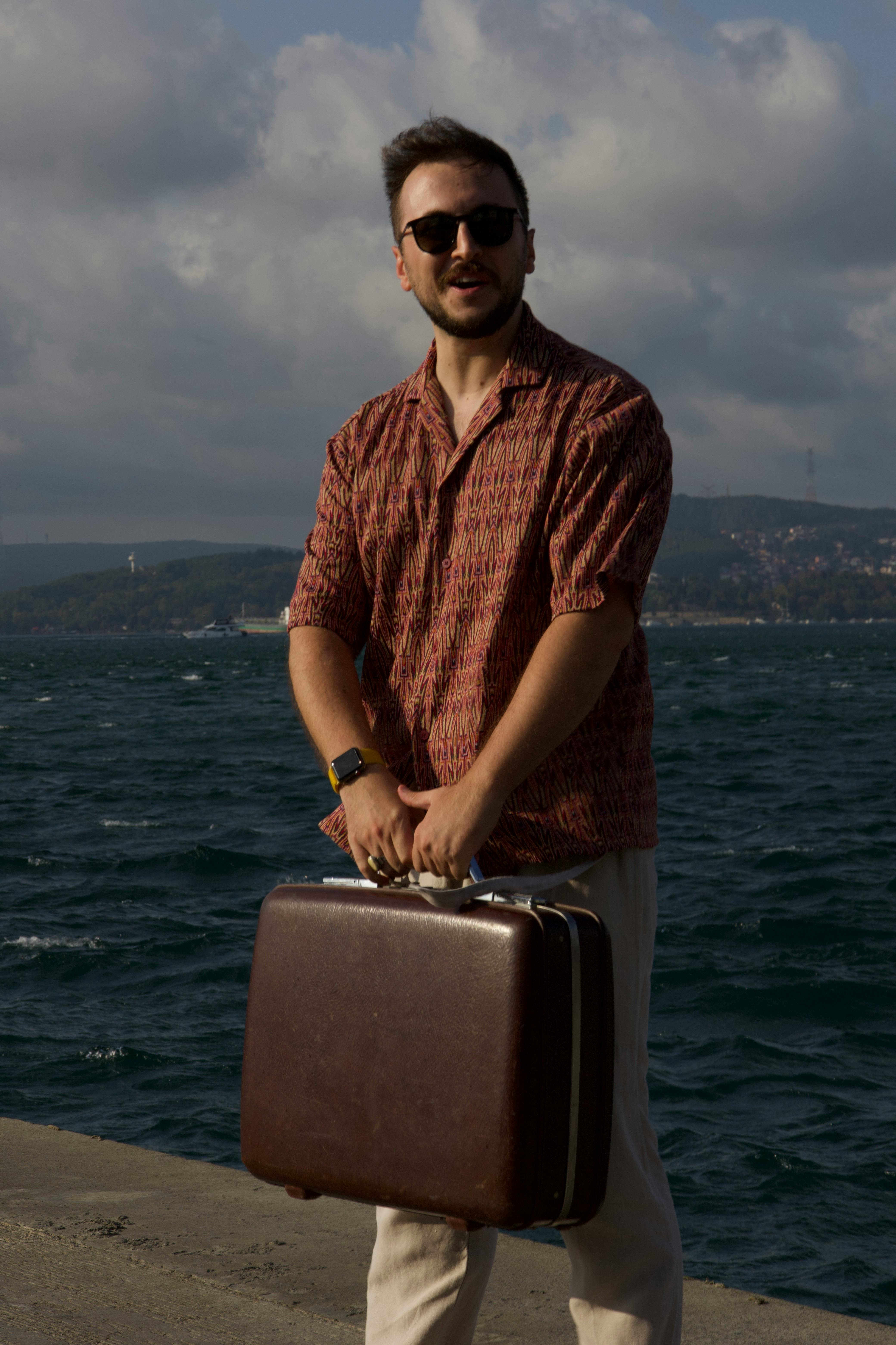 Man standing by the sea holding a suitcase, wearing sunglasses and a summer shirt.