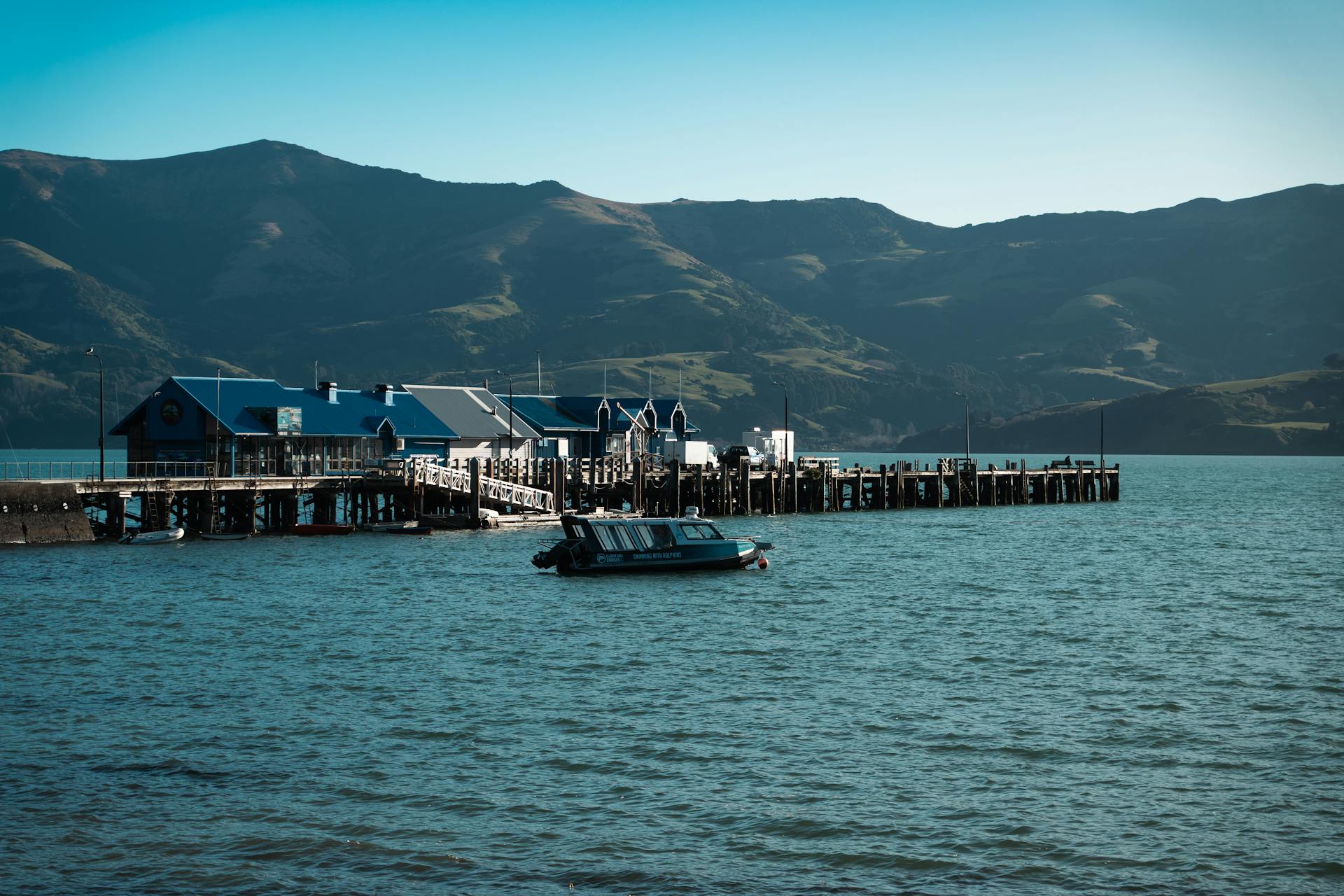 Breathtaking view of Akaroa wharf, cabins, and surrounding mountains on a clear summer day.