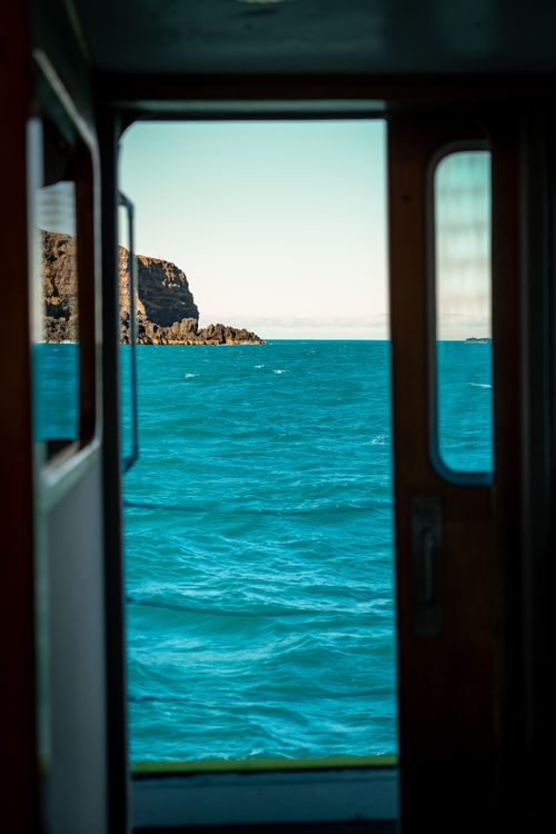 Free Scenic ocean view framed by a boat cabin's doorway, capturing tranquil blue seas and rocky coast. Stock Photo