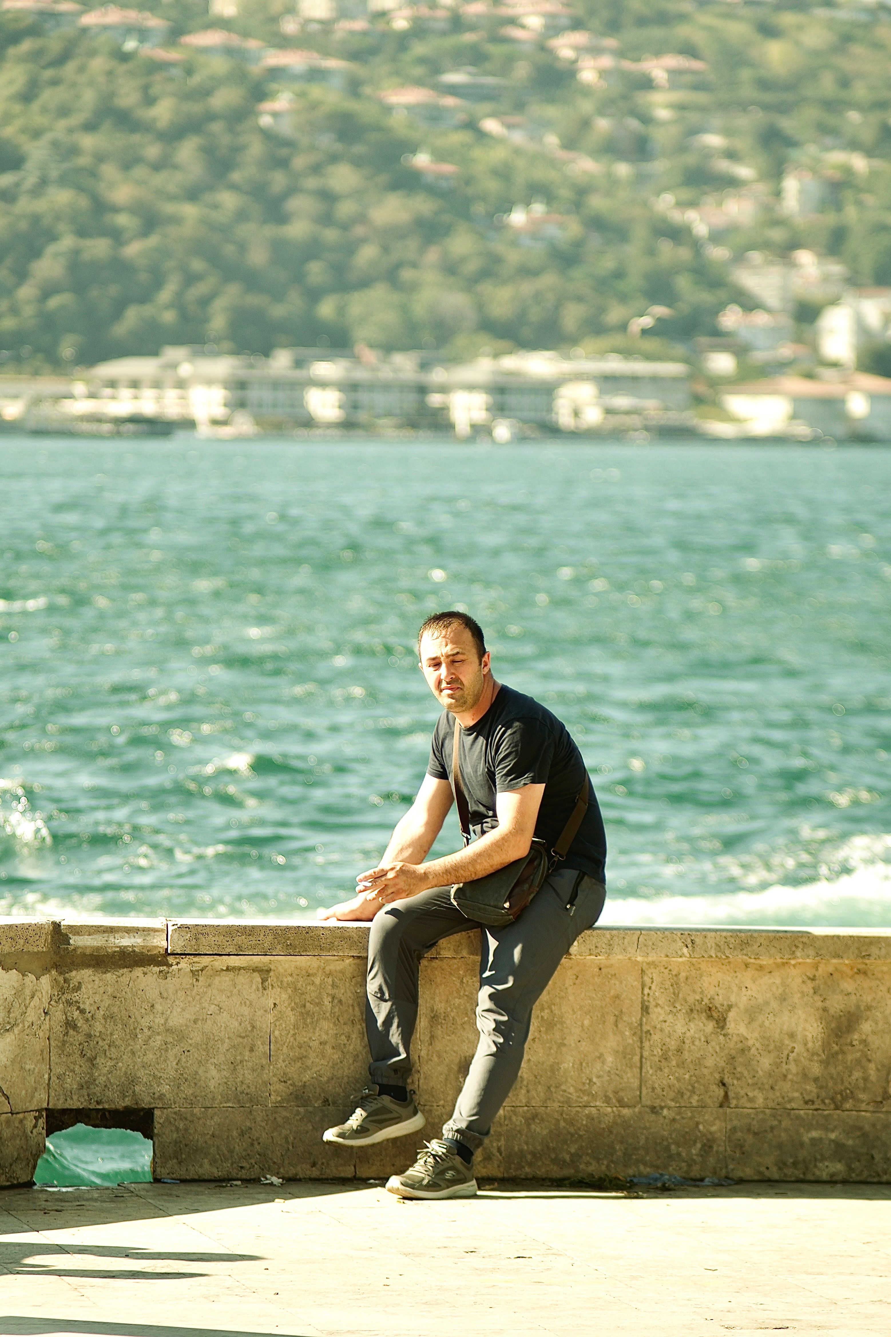 A man sits on a stone wall by the ocean, enjoying a sunny day outdoors.