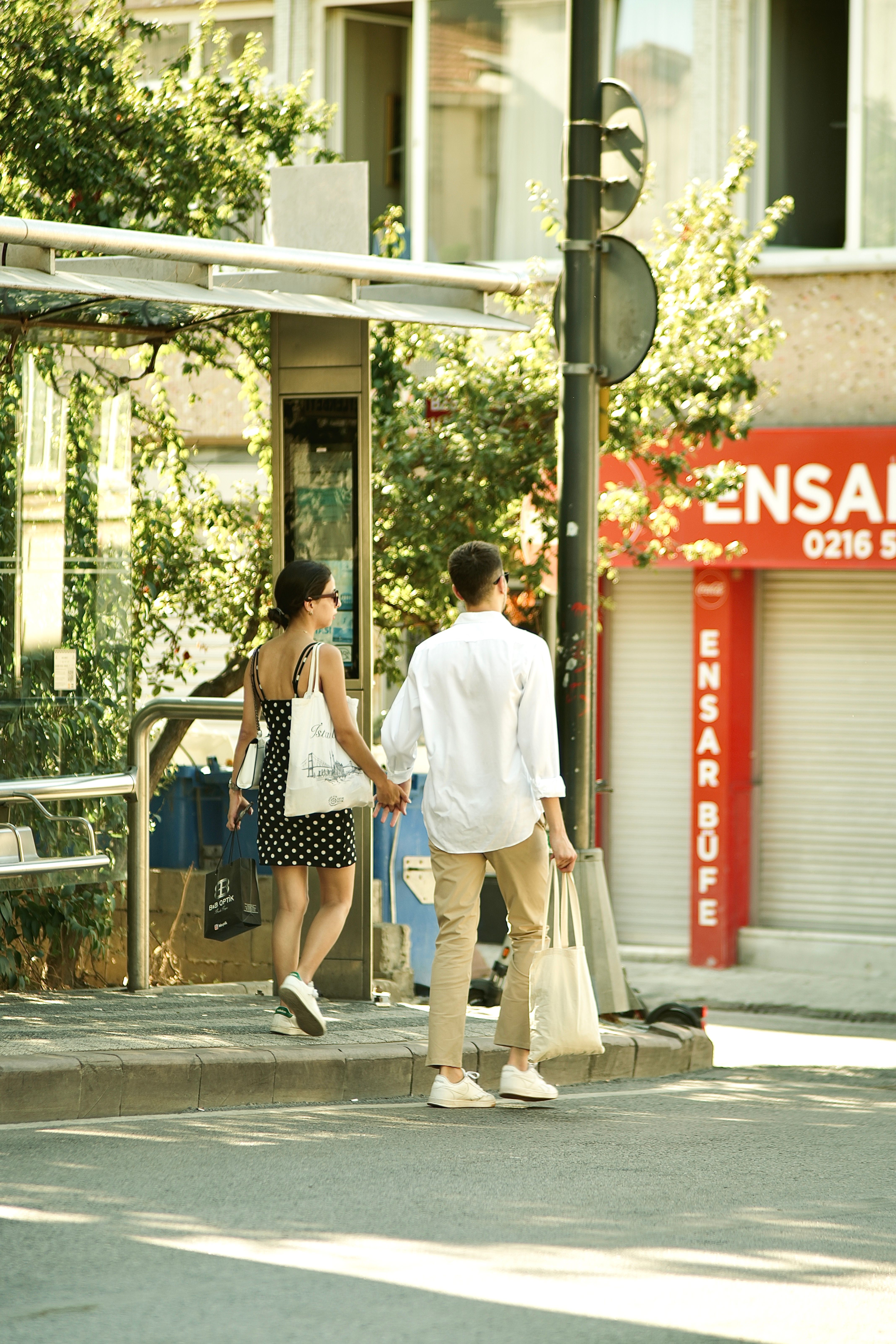 A couple holding hands while standing at a city bus stop, enjoying a sunny day.