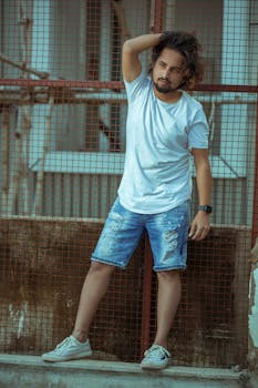 A man with long hair and beard casually poses in denim shorts and white t-shirt against an urban backdrop.
